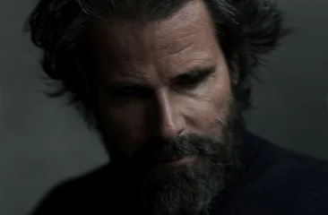 A man with a full beard and messy hair looks down.